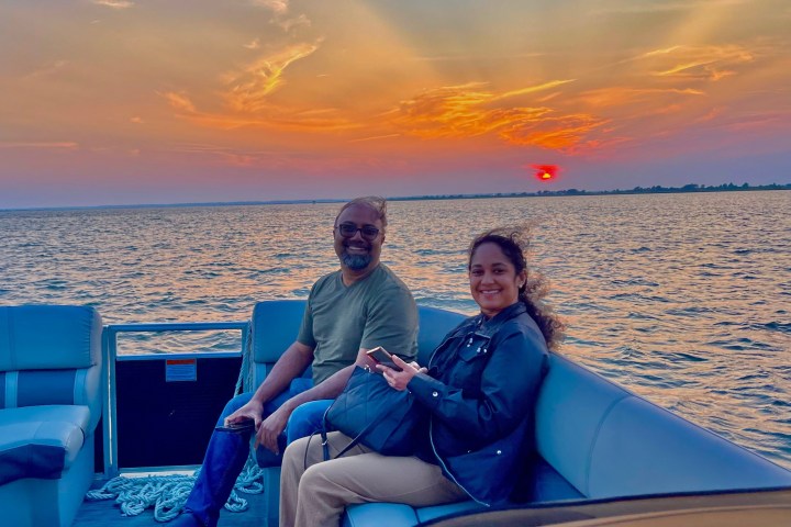 Two people smiling on a boat at sunset over calm water.