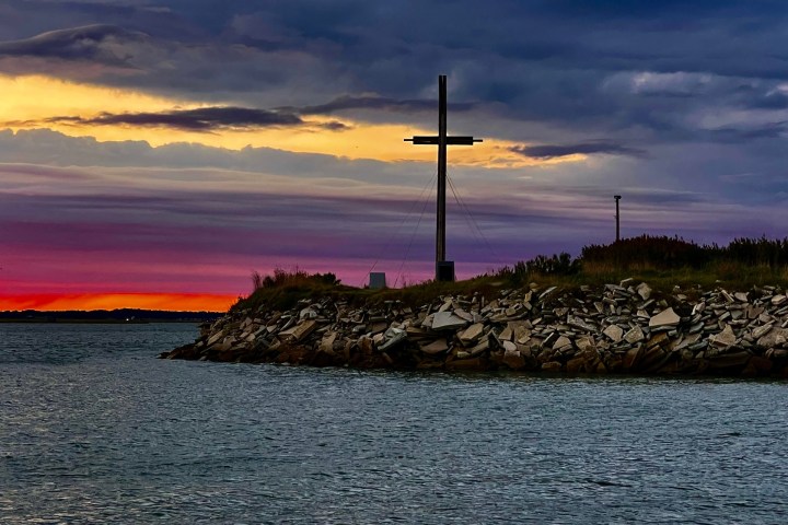 Silhouette of cross on rocky shore at sunset with cloudy sky and vibrant horizon.
