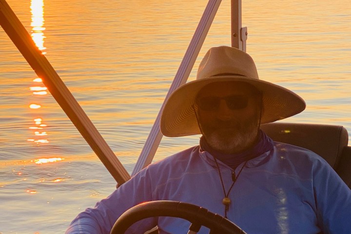 Man with hat and sunglasses steering boat at sunset on calm water.