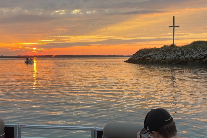 Boat on water at sunset, person sitting, cross on shoreline.