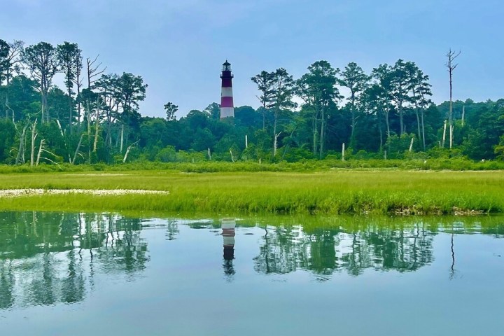 Lighthouse with trees reflected in calm water on a cloudy day.