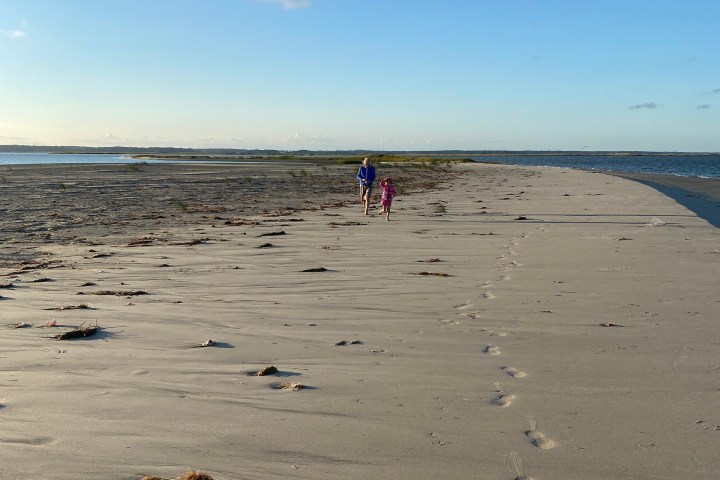 Two people walk on a sandy beach with footprints under a clear blue sky.
