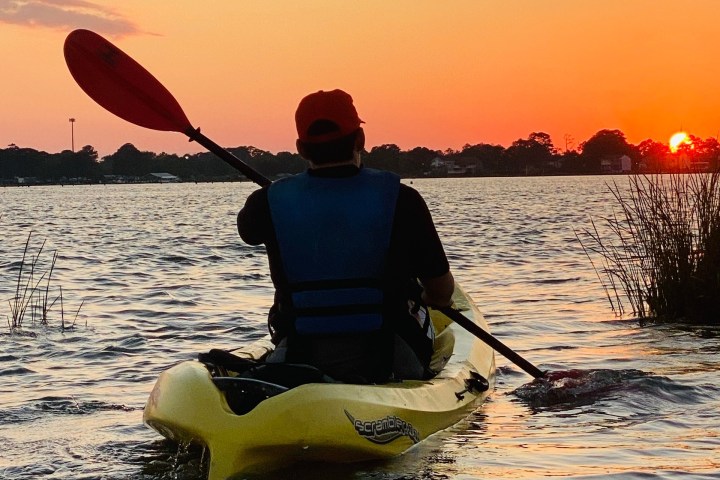 Person kayaking on a lake at sunset, wearing a red hat and blue life jacket.