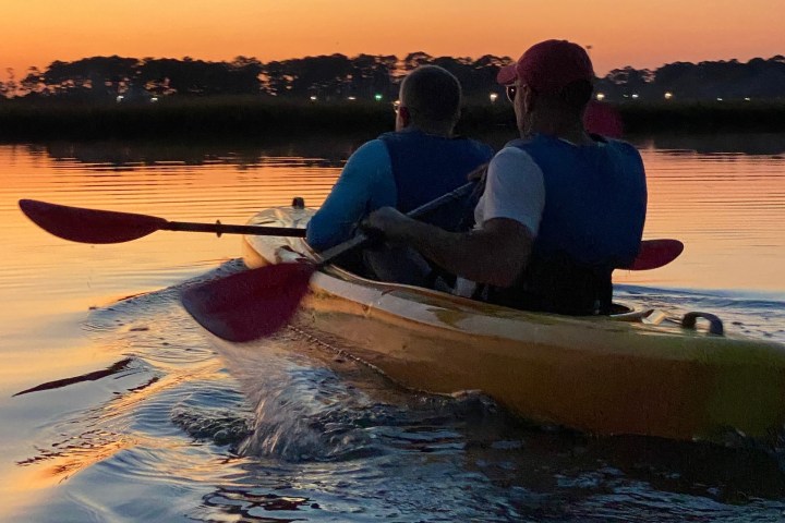 Two people kayaking on calm water at sunset with trees in the background.