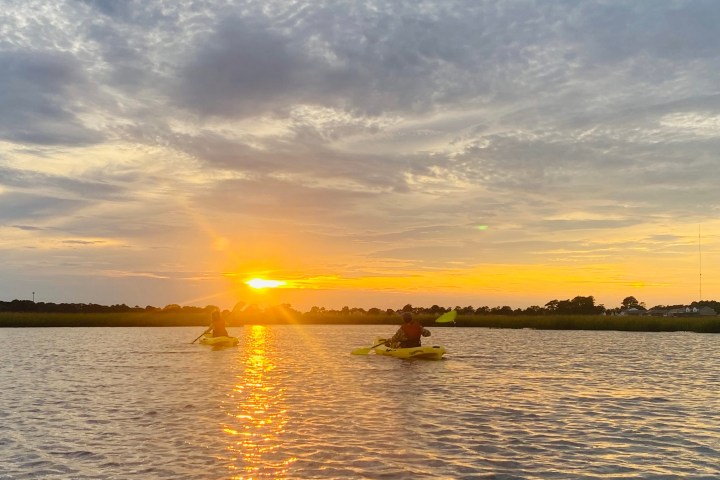 Two kayakers on water during sunset with cloudy sky.